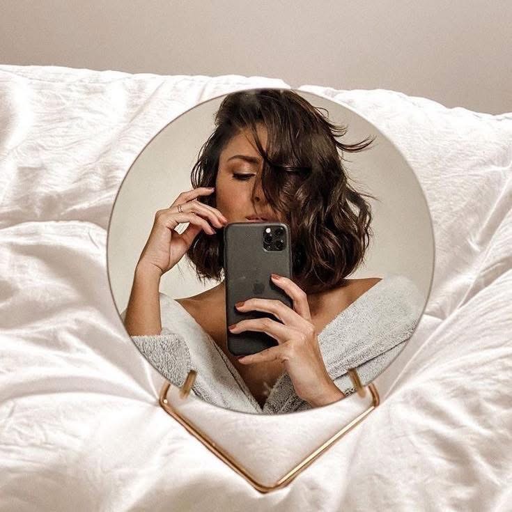 Woman taking a mirror selfie with a smartphone while sitting on a bed in a robe.